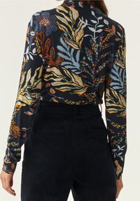 Kvinna i en långärmad marinblå blommig blus som är instoppad i högmidjade mörka byxor, sedd bakifrån med avslappnad hållning.