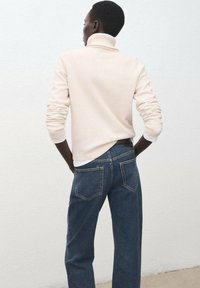 Hellrosa Rollkragenpullover mit geripptem Kragen und Bündchen, kombiniert mit dunkelblaue Jeans im entspannten Schnitt mit Gesäßtaschen.