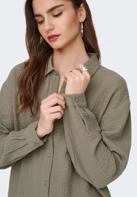 Olijfgroene blouse van gestructureerde stof, met een knoopsluiting, lange mouwen en een kraag met een losse pasvorm. Accent van een zilveren ring.