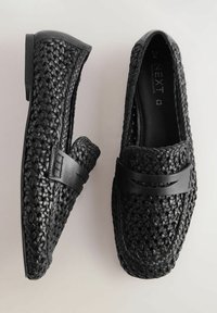 Mocasines negros trenzados con una parte superior texturizada y una banda de cuero suave a lo largo del empeine. Suela plana de goma, diseño de punta redondeada.