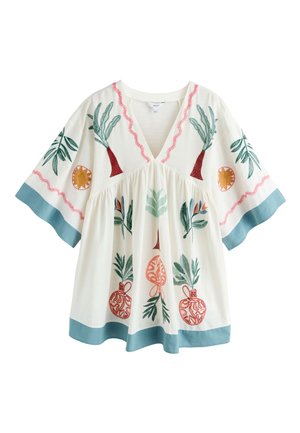 Jeune femme portant une robe blanche ample avec des imprimés colorés de plantes et de vases, debout sous un toit de chaume dans un environnement extérieur ensoleillé.