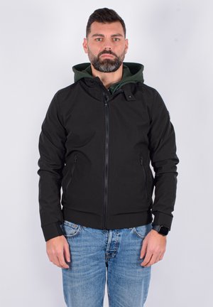 Giacca nera con zip e fodera verde a cappuccio, dotata di due tasche laterali e polsini elasticizzati. Indossata con jeans azzurri chiari.