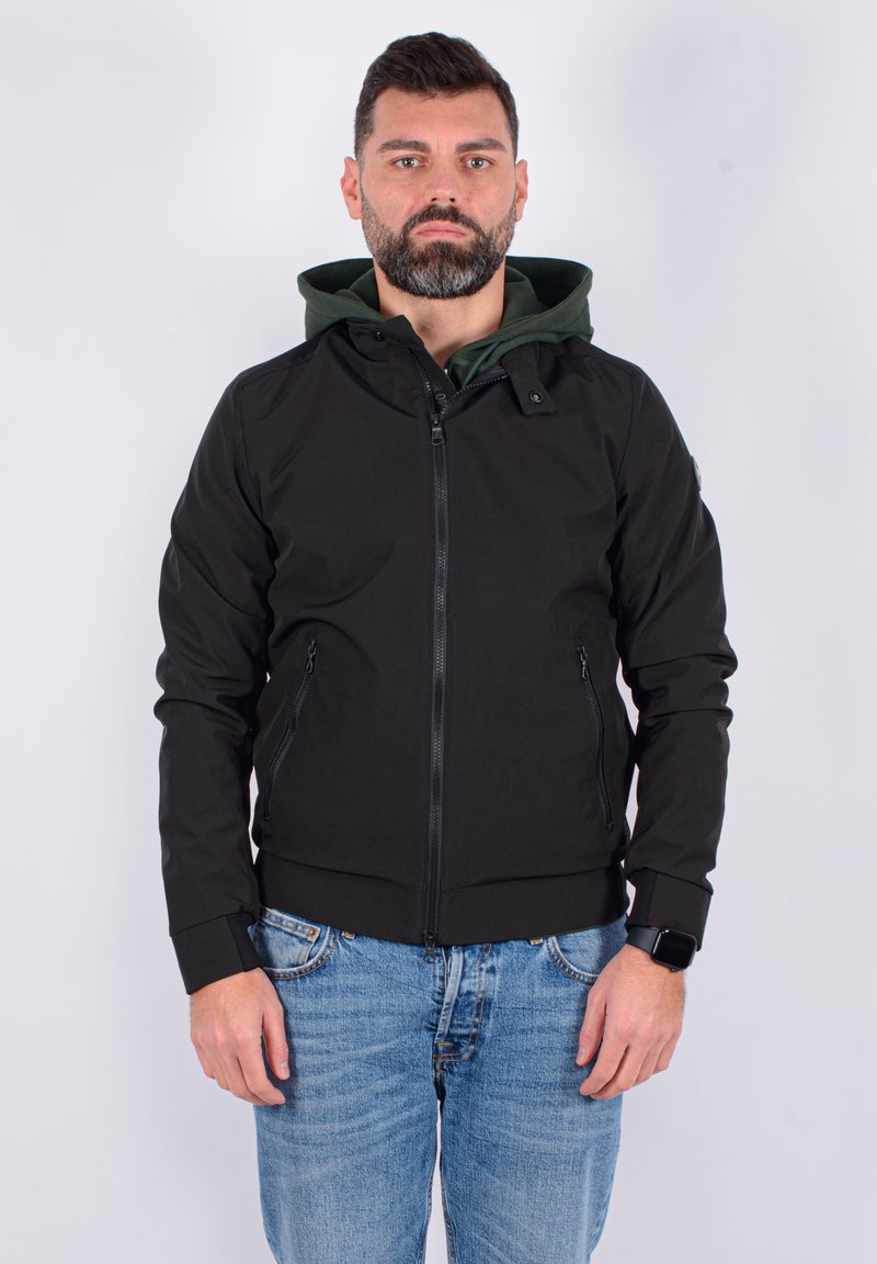 Giacca nera con zip e fodera verde a cappuccio, dotata di due tasche laterali e polsini elasticizzati. Indossata con jeans azzurri chiari.