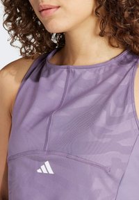 Kvindelig iført en lila ærmeløs atletisk top med subtilt camouflagemønster og hvidt Adidas-logo på fronten.