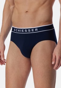 Männlicher Oberkörper, der marineblaue Unterhosen mit weißem Bund trägt, auf dem "SCHIESSER"-Markierung steht, vor einem schlichten weißen Hintergrund stehend.