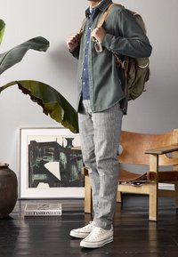 Persoon in grijs geblokte broek, witte sneakers, denim shirt, groene overshirt en met een olijfgroene rugzak in een kamer met planten en kunstwerken.
