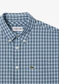 Camisa de cuadros azules con cuello, hecha de tela, que presenta botones blancos y un logo bordado verde en el lado izquierdo.