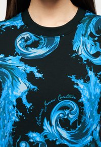 Μαύρο μπλουζάκι με στρογγυλή λαιμόκοψη και μοτίβα με περιστρεφόμενα μπλε νερά, με το κείμενο «Versace Jeans Couture» κοντά στο κέντρο του στήθους.