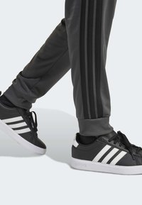 Zapatillas negras de Adidas con tres franjas blancas, combinadas con pantalones deportivos gris oscuro que tienen franjas laterales negras y puños acanalados.