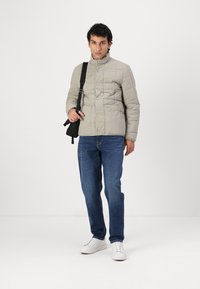 Világosszürke steppelt dzseki magas gallérral és gombos záródással, kék farmernadrággal és fehér sportcipővel párosítva. A modellen fekete válltáska van.
