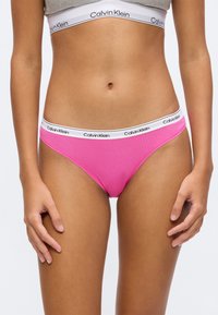 Femme portant des sous-vêtements Calvin Klein roses avec une ceinture blanche griffée et un soutien-gorge assorti, montrant la taille et les mains détendues.