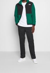 Fleece bunda se zelenými a černými panely, zipovými kapsami, v kombinaci se šedými kalhotami a bílými teniskami. Jednoduchý design a současný střih.