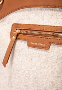 Borsa in tela color beige chiaro con superficie testurizzata, arricchita da una rifinitura in pelle marrone e una cerniera color oro con un'etichetta che recita "GERRY WEBER."