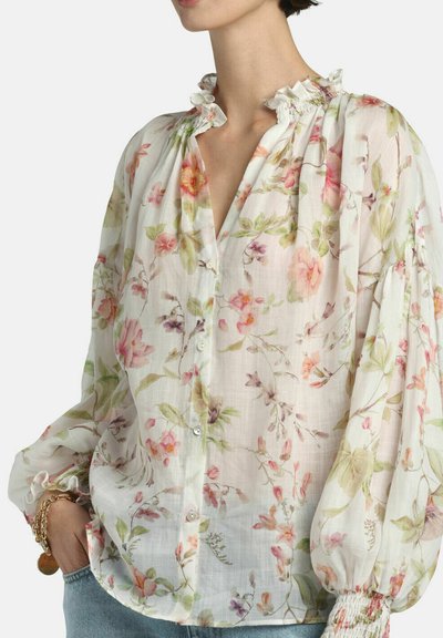 Kvinde iført en gennemsigtig hvid bluse med blomsterprint, flæsekant og puffede lange ærmer, kombineret med lyseblå jeans og gyldne armbånd.