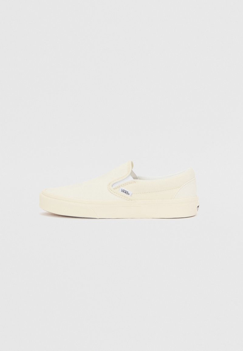 Adidași slip-on din pânză crem cu o suprafață texturată, talpă din cauciuc, accente elastice pe laterale și un mic etichetă de brand pe partea exterioară.
