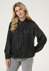 Blusa de satén negro con cuello alto y volantes, tapeta frontal y detalles de botones. Mangas largas con puños fruncidos. Textura suave.