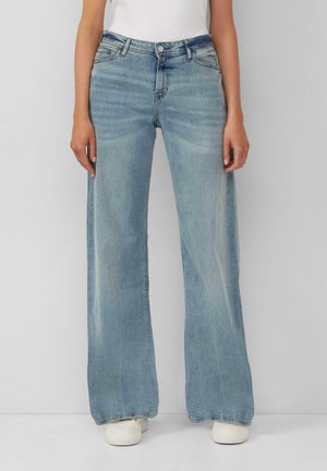 Femme portant un jean large bleu clair, des baskets blanches et un haut blanc, debout devant un fond gris uni.