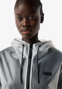 Close-up of a person wearing a white translucent rain jacket with black drawstrings and a "Hunter" logo patch on the chest.