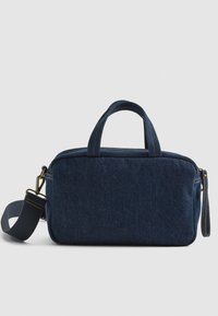 Bolso de denim azul oscuro con dos asas cortas, una correa ajustable y cierre de cremallera. Presenta costuras amarillas y una textura suave.
