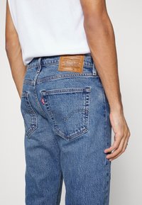 Blå denimjeans med en blekt textur, rak passform och dubbla bakfickor, med en brun läderlogga i midjan.