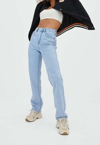 Persona que lleva jeans de tiro alto color azul claro, zapatillas beige, top corto blanco y chaqueta negra levantada sobre los hombros, contra un fondo blanco.