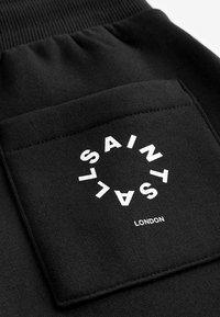 Sort sweatstof med en tekstureret lomme, der har en hvid cirkulær logo-tekst "SAINT L" over "LONDON" nederst.
