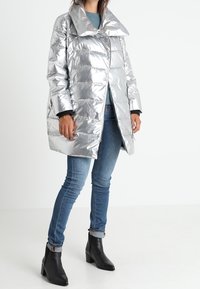 Srebrna puchowa kurtka z dużym kołnierzem, zapięciem na zamek błyskawiczny i błyszczącą, gładką teksturą. Połączona z niebieskimi dżinsami i czarnymi botkami do kostki.