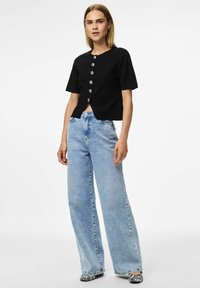 Melnā cropped adīta blūze ar īsām piedurknēm un sudraba pogu detaļām, apvienota ar gaiši ziliem platām biksēm un melnbaltiem rakstainiem apaviem.