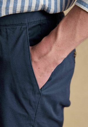 Adult’s hand resting inside the front pocket of navy blue pants, wearing a blue and white striped shirt.