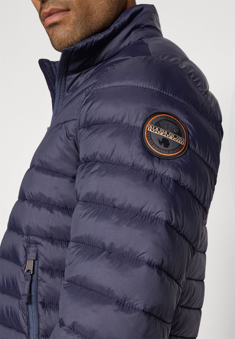 Marineblaue Steppjacke mit hohem Kragen, gesteppter Textur und einem runden Logo-Patch auf der linken Schulter. Seitlicher Reißverschlussdetail sichtbar.