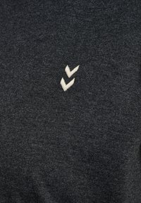 Donkergrijs stof met een zachte, textuurafwerking; heeft een wit geborduurd logo van twee gestapelde chevrons aan de linkerkant.