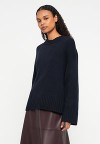 Žena s kovrčavom kosom koja nosi tamnoplavi pleteni pulover i smeštnu kožnu suknju, stoji ispred jednostavne bele pozadine.