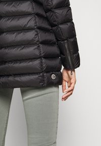 Černá péřová bunda s prošívanými sekcemi, na rukávu se zipem a logem u lemu. Nošena přes světle zelené džíny.