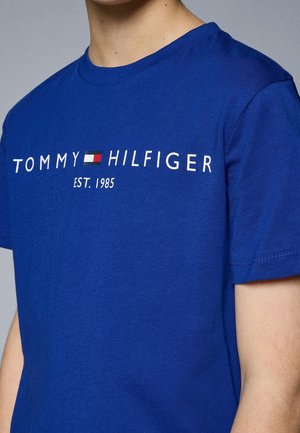 Person iført en blå Tommy Hilfiger T-shirt med hvid tekst og et lille rødt, hvidt og blåt flaglogo, teksten lyder "EST. 1985."