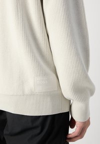 Maglione lavorato a maglia di colore chiaro con una trama a coste, maniche lunghe e un'etichetta opaca con la scritta "BETTER BOSS" sul lato inferiore.
