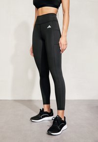 Czarne legginsy z wysokim stanem o teksturowanym wykończeniu, z logo Adidas na lewej udzie oraz płaskimi szwami dla wygody. Czarne buty sportowe.