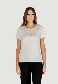 Donna con lunghi capelli castani, che indossa una maglietta bianca con grafica e pantaloni neri, su uno sfondo leggero e neutro.