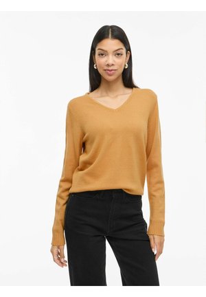 Femme aux longs cheveux foncés portant des boucles d'oreilles en anneau dorées, un pull moutarde à col en V et un pantalon noir, debout sur un fond blanc.