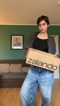 Pappkartong märkt "zalando" hålls av en person i svart topp och överstor blå jeans. Bakgrunden inkluderar en grön vägg och en soffa.