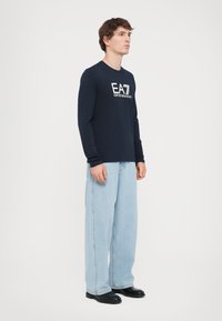 Marineblå langermet skjorte med svart "EA7 EMPORIO ARMANI" logo, brukt med lyseblå vide jeans og svarte sko. Enkelt, uformelt design.