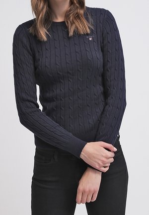 Kvinde iført en marineblå kabelstrikket sweater med lange ærmer og sorte bukser, stående med armene krydset ved taljen.