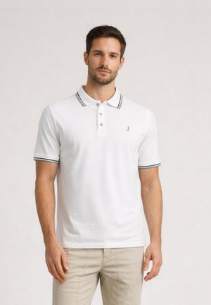 Uomo che indossa una maglietta polo bianca a maniche corte con rifiniture nere sul colletto e sulle maniche, abbinata a pantaloni beige, in piedi davanti a uno sfondo semplice.