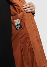 Bruine binnenvoering van een jas, met een geweven logopatch en een waslabel, die een gladde textuur en eenvoudige designelementen toont.