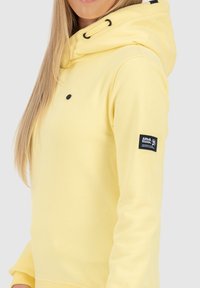 Vrouw in een lichtgele hoodie met een zwart etiket op de mouw en lange blonde haren zichtbaar.