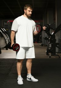 Man in een loszittend wit T-shirt en shorts houdt zwarte dumbbells vast met rode labels. Sportschoolomgeving met oefenapparatuur op de achtergrond.