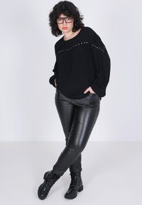 Zwarte blouse met lange mouwen en studs; gecombineerd met glanzende zwarte nepleer legging en zwarte veterschoenen. Effen witte achtergrond.