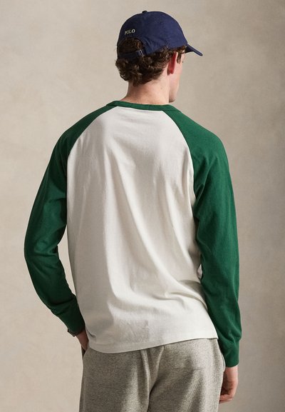 Άνδρας που φοράει λευκό πουκάμισο με μακριά μανίκια και πράσινα ραγκλάν μανίκια, γκρι παντελόνια και μπλε σκούφο Polo, στραμμένος με την πλάτη στην κάμερα.