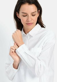 Chemise blanche à boutons, avec col, manches longues, poche poitrine et détails à rabat. Le tissu semble lisse et légèrement texturé.