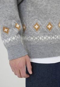 Pull gris tricoté avec un poignet côtelé, présentant des motifs en losange orange et blanc. Porté par-dessus une chemise blanche, montré avec une main.