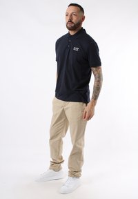 Polo navy con cuello y logo de EA7, combinado con pantalones beige y zapatillas blancas. El tejido parece suave y ligero.